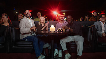 Keegan-Michael Key, Chelsea Handler, Kumail Nanjiani, and Kevin Hart seated at a round table in a dimly lit comedy club or lounge, smiling and watching a show, with a glowing candle on the table and an audience in the background.