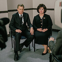 A man and woman in formal black attire sit side by side in a plain office-style room, holding hands, looking serious, as they are being interviewed by someone off camera, with a video camera visible in the foreground.