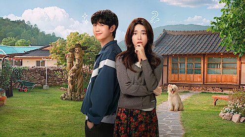 A young man and woman stand in a sunny, rural yard with traditional wooden house, lawn, garden statues, and a fluffy dog, mountains in the background, blue sky with clouds, both subjects displaying thoughtful and playful expressions.