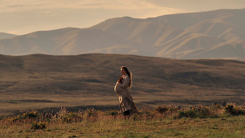 Pregnant woman in a long dress stands in a grassy field with wildflowers, set against a backdrop of rolling hills and mountains during golden hour, creating a serene and natural outdoor scene.