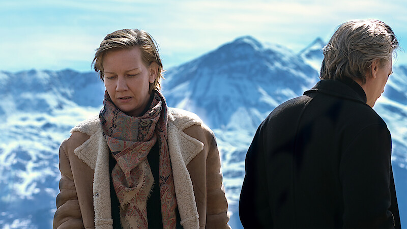Two people stand outdoors in winter clothing with snowy mountains in the background, one facing forward and the other with their back turned, suggesting a somber or reflective mood in a cold, alpine environment.