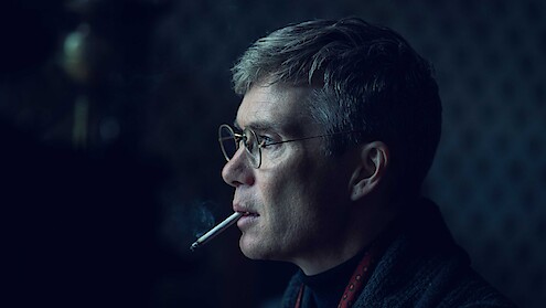 A man with short hair and glasses in a dark, softly lit room is shown in profile view, wearing a dark jacket and smoking a cigarette, smoke rising in front of his face, with a blurred, patterned background.