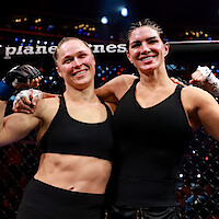 Two female mixed martial artists in black sportswear smiling and posing together inside a cage after a match, arena lights and audience visible in the background.