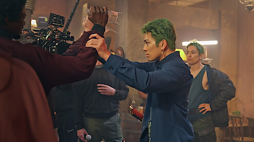 A film crew shoots an action scene indoors with a man with green hair in a blue outfit rehearsing martial arts choreography, while cameras and crew members observe in a dimly lit, industrial set.