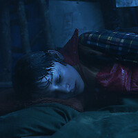 A young boy in a plaid shirt and red vest lies curled up on his side in a dimly lit setting, appearing wet, alone, and cold.