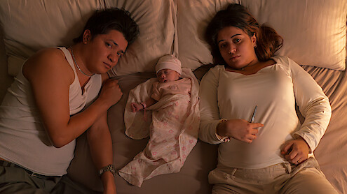 A man, woman, and newborn baby lie in bed together, parents on either side of the baby who is swaddled and wearing a hat. The mood is subdued, with soft, warm lighting suggesting nighttime or early morning at home.