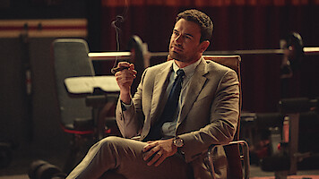 Man in a tan suit sitting in a chair indoors, holding a cigar and looking thoughtful. The background features gym or fitness equipment and dim, moody lighting.