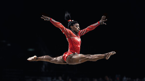 Gymnast Simone Biles doing a split leap in the air, wearing a red leotard.
