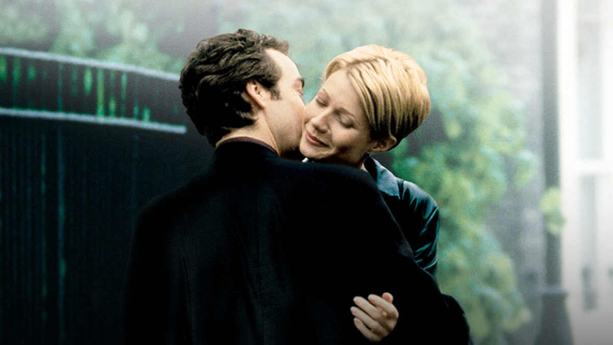 A man and woman embrace outdoors near a garden gate, with greenery and soft lighting in the background, conveying a romantic and intimate atmosphere.