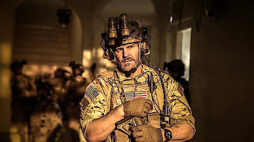 Armed soldier in camouflage gear and helmet with night vision goggles stands in a dimly lit hallway, American flag patch visible, with other soldiers blurred in the background.