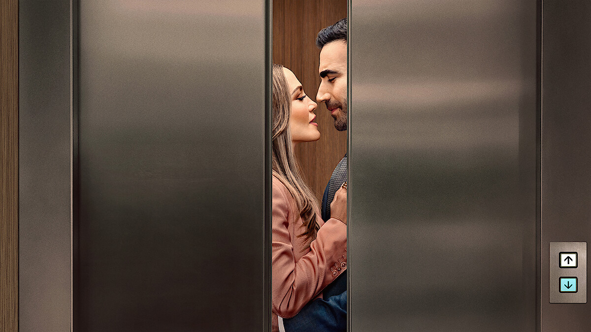 A man and woman standing closely, facing each other romantically inside an elevator as the doors begin to close, suggesting a private or intimate moment in a modern building setting.