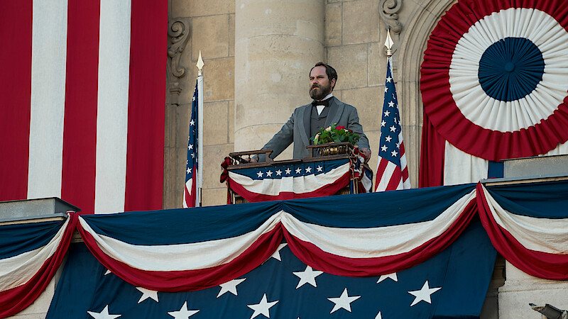 A man in historical attire stands on a decorated balcony with American flags and patriotic bunting, delivering a speech in a formal, historic setting. The mood is solemn and ceremonial.