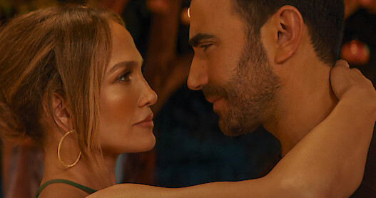 Jennifer Lopez as Jackie Cruz and Brett Goldstein as Daniel Blanchflower facing each other closely, embracing in a romantic setting with warm lighting and a blurred background, suggesting an intimate or emotional moment.