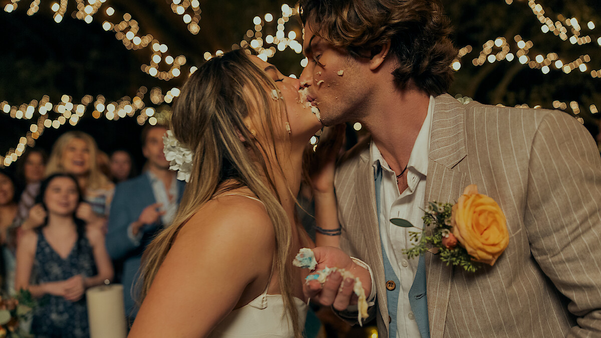 Bride and groom with cake on their faces share a kiss at an outdoor wedding reception. String lights create a festive atmosphere, while guests watch in the background. The groom wears a flower boutonniere.