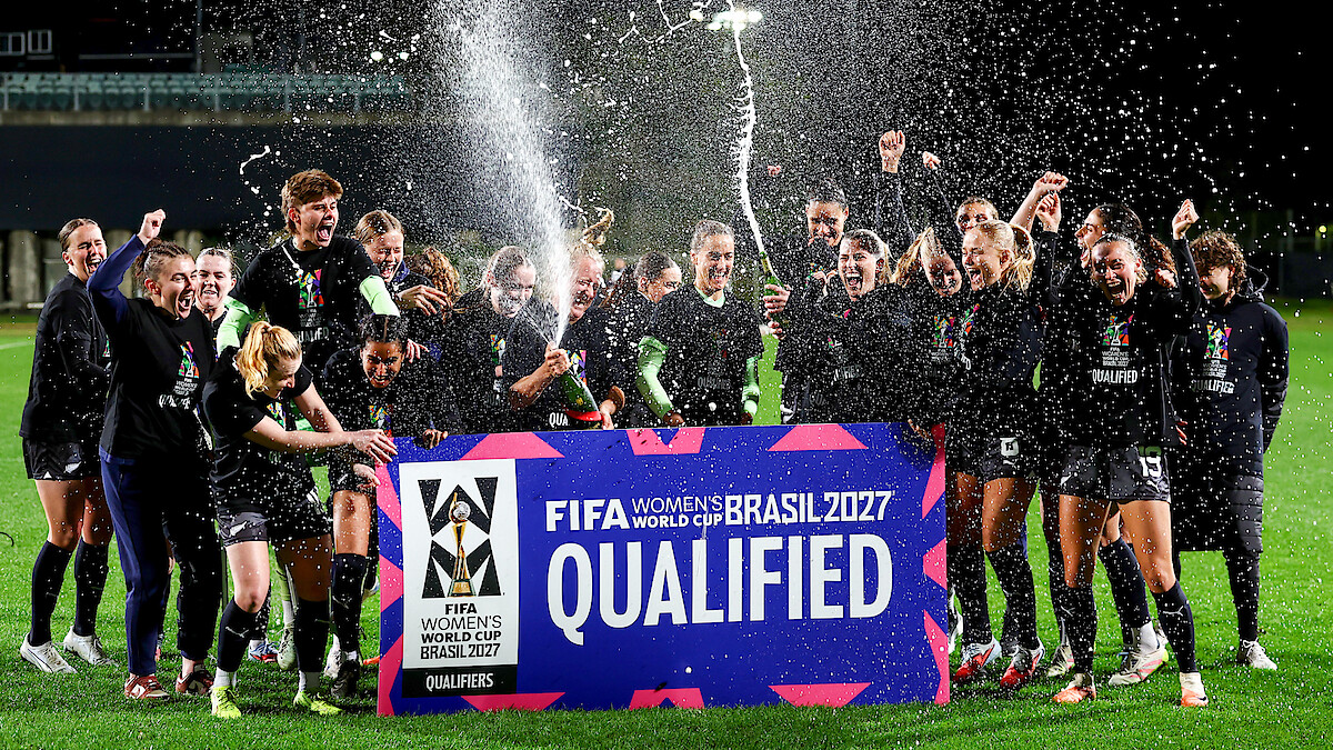 Women's soccer team celebrates at night on a field, spraying champagne and cheering behind a large sign reading "FIFA Women's World Cup Brasil 2027 Qualified." Bright stadium lights and festive atmosphere.