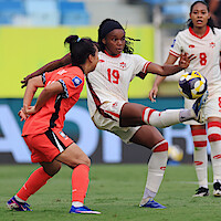 Soccer match action shot with a player in a white and red uniform kicking a ball while a player in an orange uniform attempts to block; outdoor stadium, other players and audience blurred in background.
