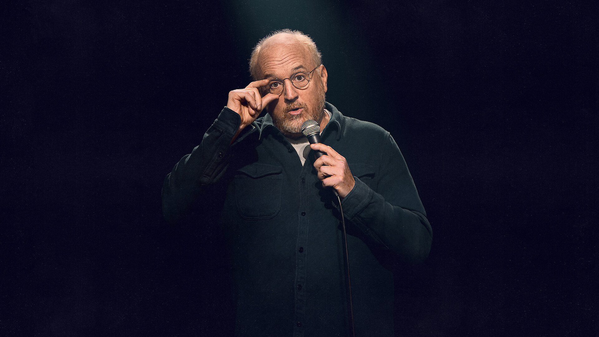Middle-aged man with glasses performs stand-up comedy on stage, holding a microphone under a spotlight and speaking to the audience, with a dark, simple background.