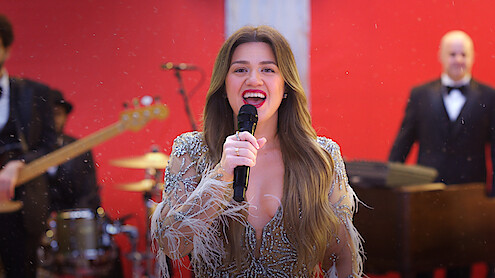 Woman in a sparkling dress sings into a microphone on stage with a band, red background, festive and lively atmosphere.