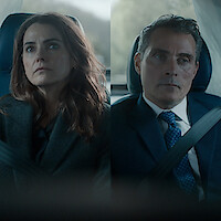 Two people, a woman and a man, sit side by side in a car wearing seatbelts. Both appear serious and are dressed in business attire, with a blurred background visible through the car windows.