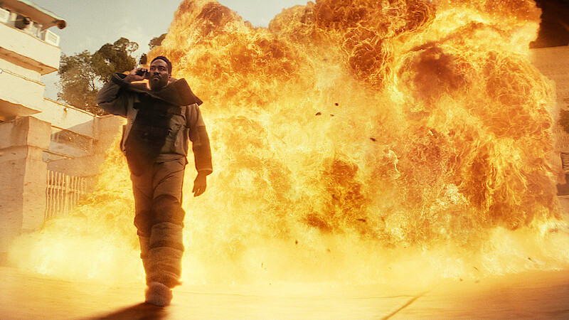 A person in tactical gear walks confidently away from a massive explosion in an urban environment, with flames and debris filling the background.
