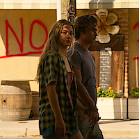 Two young people walking outside in daylight, with a woman in the foreground and a man beside her. They are near a building with a large red "NO" graffiti, decorated flower, wooden barrels, and a handwritten chalkboard sign.