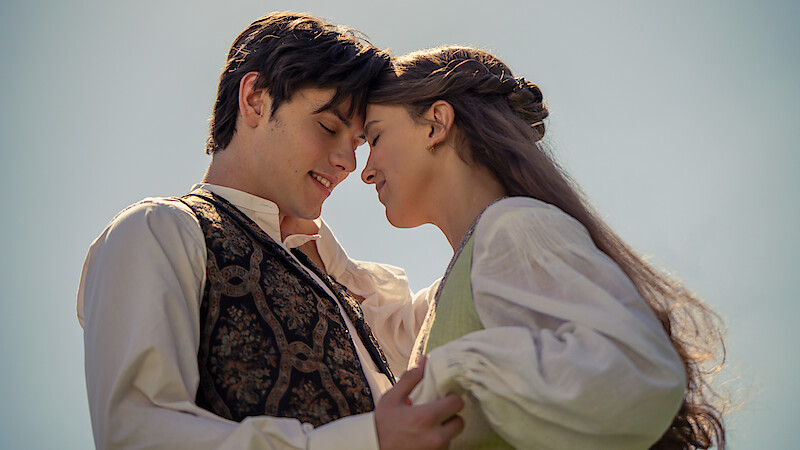 Young couple in vintage clothing embracing outdoors on a sunny day, touching foreheads and smiling with affection, clear sky in the background.