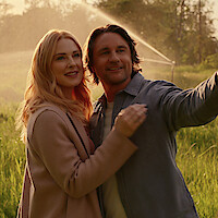 A smiling couple standing together in a sunlit field, surrounded by green grass and trees, with a water sprinkler in the background, enjoying a peaceful outdoor moment.