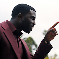 Man in a burgundy suit pointing and speaking with intensity outdoors, with blurred trees and cloudy sky in the background.