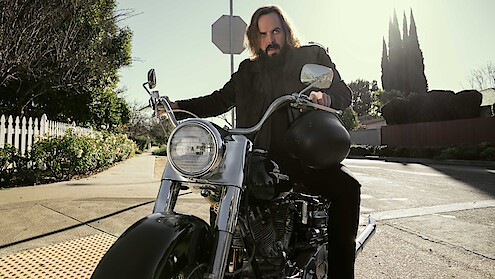 Man with long hair and beard sitting on a motorcycle in a suburban neighborhood, holding a helmet, with trees, a white picket fence, and stop sign visible in the sunlit background.