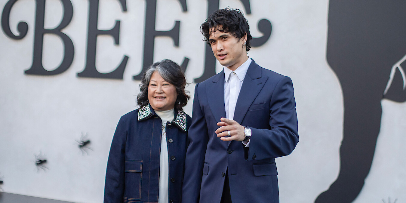 Two people stand together, smiling, in front of a wall with large black text and silhouettes as part of an event backdrop. The man wears a dark suit and white shirt, while the woman wears a dark jacket with a light collar.