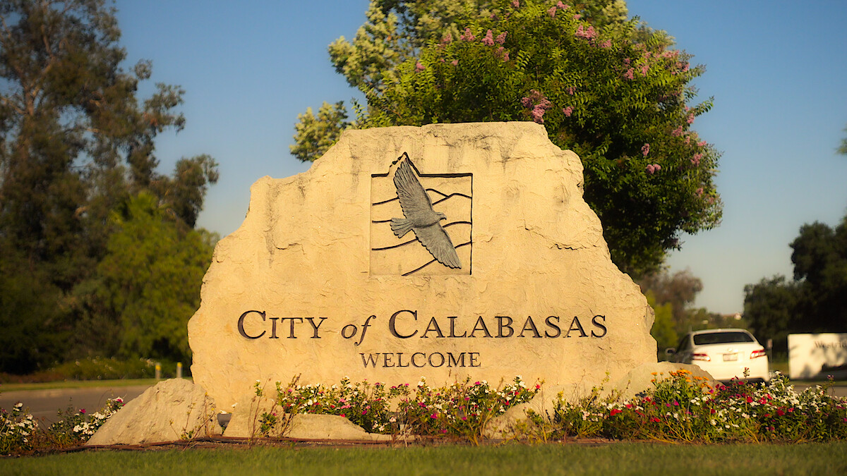 Large stone welcome sign for the City of Calabasas with an eagle engraving, surrounded by flowers, trees, and parked cars in a sunny outdoor setting.
