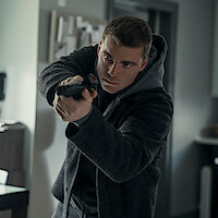 Serious man in a dark coat and hoodie aiming a handgun indoors, standing in a tense posture inside a modern, dimly lit apartment or office environment.