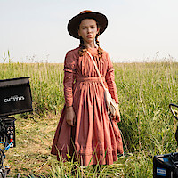 Young girl in a pioneer dress and hat standing in a grassy field on a film set, with camera equipment visible around her on a bright day.
