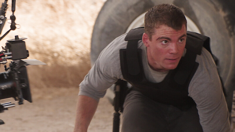 Man in tactical gear crouches on a sandy set beside large tires, with a professional film camera visible, indicating a movie or TV production in a rugged outdoor environment.