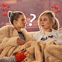 Joanne (Kristen Bell) and Morgan (Justine Lupe) sit close together under a blanket on a couch, looking at each other with curiosity and affection, surrounded by heart icons and a question mark. Cozy and warm atmosphere.