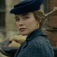 Woman in vintage blue clothing and hat looks over her shoulder outdoors, with a blurred background featuring sacks and bricks, suggesting a historical or period setting.