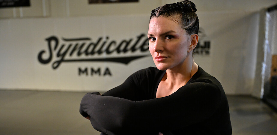 Gina Carano with braided hair in athletic wear sits with arms crossed in an MMA gym, "Syndicate MMA" visible on the wall behind her, suggesting a focused training environment.
