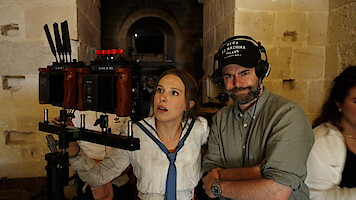 A woman in costume and a man with headphones stand by a camera rig in a stone hallway on a film set, reviewing footage together.
