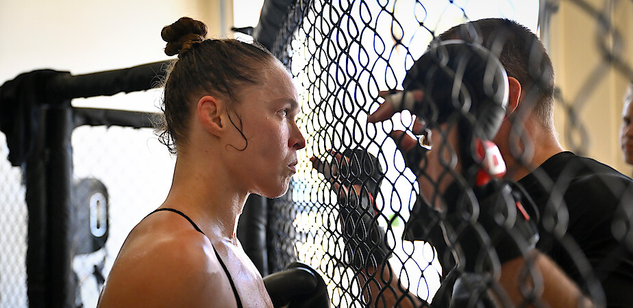 Ronda Rousey listens to coach’s advice beside cage during training or a match, sweat on her face, intense focus, gym environment, daylight streaming in, black gloves and athletic attire, supportive coaching moment.