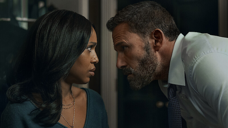 A serious man in a dress shirt and tie leans in close to a concerned woman indoors, their faces inches apart, suggesting tension or intense conversation in a dimly lit room.