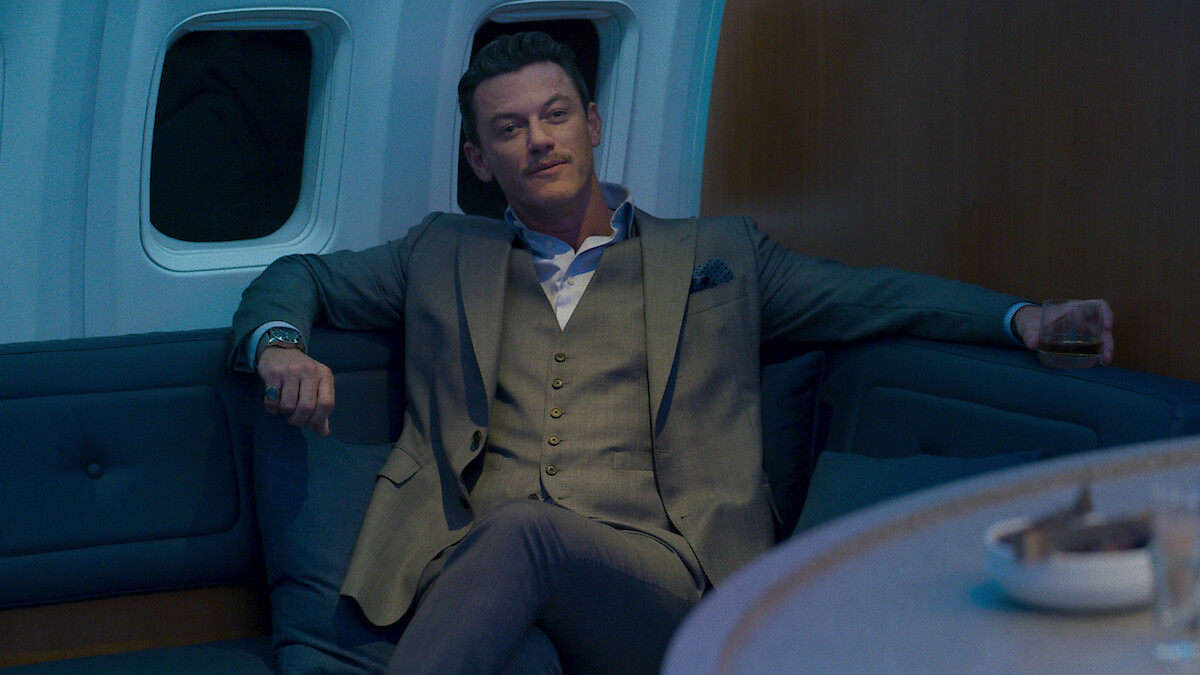 A man in a suit sits relaxed on a blue sofa inside a plane cabin, holding a drink with an arm resting on the seat, in a dimly lit, modern and luxurious environment.