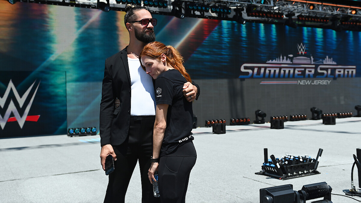 Two people embrace in a staged outdoor arena with SummerSlam and WWE logos in the background, under bright sunlight, conveying a calm and supportive mood before a wrestling event.