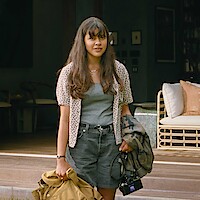 Young woman with long hair stands outside a cozy, modern house holding a coat, bag, and camera. The relaxed setting features a wicker sofa and bookshelves, evoking a calm, casual, and welcoming atmosphere.