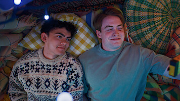 Two young men lying on colorful patterned blankets, one in a knit sweater and the other in a blue shirt, surrounded by pillows and soft lighting, creating a cozy, intimate indoor setting.