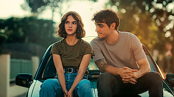A young woman and man sit on the hood of a car outdoors, surrounded by trees and soft sunlight, appearing relaxed and engaged in conversation in a quiet suburban setting.