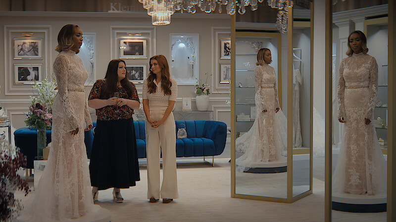 Three women in a stylish bridal shop, one wearing a wedding gown standing by mirrors, with elegant decor, framed photos on the wall, a blue couch, and chandeliers creating a classy atmosphere.