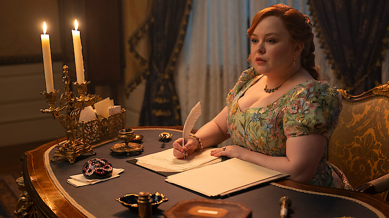 Woman in a floral dress writes with a quill at an ornate wooden desk with candles, in a lavish, candlelit room with elegant curtains and antique decor.