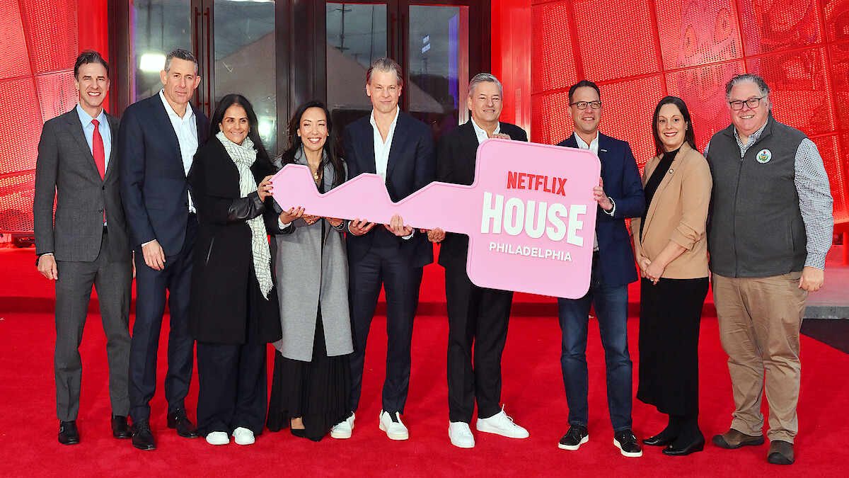 A group of people stand smiling on a red carpet in front of a building, holding a large pink key with "Netflix House Philadelphia" written on it, celebrating an event in a festive, welcoming atmosphere.