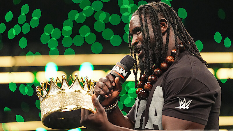 Wrestler with long braids and beaded necklace speaks into a microphone labeled "RAW" while holding a large golden crown, standing in a brightly lit arena with green lights in the background.