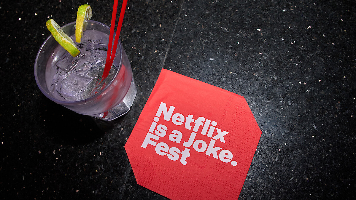 A cocktail with lime slices and two red straws sits on a black granite surface next to a red napkin that reads "Netflix is a Joke Fest" in bold white letters.